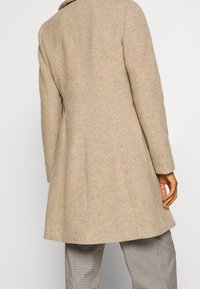 Abrigo de lana beige con una silueta ajustada, que presenta un cuello muesca, mangas largas y un dobladillo ligeramente acampanado, llevado sobre pantalones de cuadros.