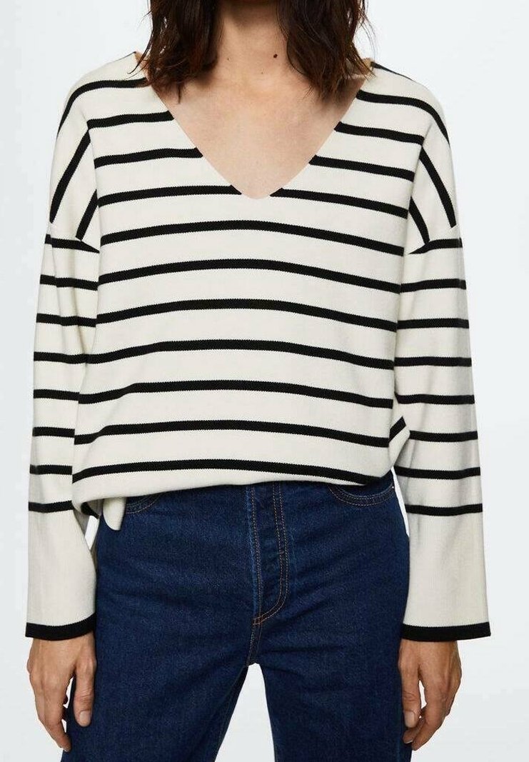 Femme portant un pull blanc à col en V avec des rayures horizontales noires et un jean bleu foncé, debout devant un fond uni.