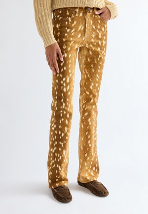 Personne portant un pull beige, un pantalon marron avec un motif tacheté blanc rappelant le pelage de cerf, et des mocassins marron, debout devant un fond uni.