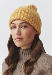 Berretto di maglia giallo con texture a coste, caratterizzato da una parte superiore leggermente appuntita. Indossato con un maglione marrone a collo alto, che mostra un aspetto accogliente.