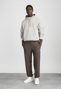 Hanorac gri deschis cu un buzunar frontal și șnururi, asortat cu pantaloni maro și adidași albi. Material neted, cu o croială lejeră.