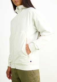 Chaqueta de cremallera color beige claro con un cuello alto, bolsillos laterales y puños acanalados, hecha de un material suave y elástico. Pequeño acento de bandera en el dobladillo.