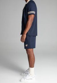 Navy short-sleeve button-up shirt with cream collar and striped sleeves, paired with navy shorts featuring a gold logo. White sneakers and socks.