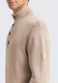 Pulover bej din tricot cu guler înalt cu nervuri, trei butoane și detalii texturate pe umeri. Prezintă o eticheta din material pe mânecă.