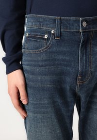 Τζιν παντελόνι σε σκούρο μπλε χρώμα με ελαφριά πλύση, διαθέτοντας πέντε τσέπες, πορτοκαλί ραφές και μεταλλικά αξεσουάρ στο μπροστινό μέρος.