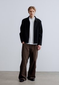 Μαύρο μάλλινο σακάκι πάνω από λευκό μπλουζάκι με στρογγυλή λαιμόκοψη, σε συνδυασμό με φαρδιά καφέ παντελόνια και μαύρα παπούτσια. Ομαλές υφές και άνετη εφαρμογή.