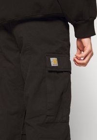 Czarne spodnie bojówki wykonane z trwałego materiału, z boczną kieszenią ozdobioną kwadratową naszywką z logo Carhartt oraz luźnym krojem.