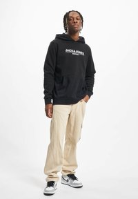 Joven de pie con pantalones cargo beige, sudadera negra con el texto "JACK & JONES" y zapatillas blancas-grises contra un fondo blanco.