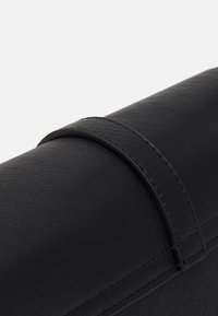 Černá kožená kabelka s texturovaným povrchem, která má zavírání s rolovaným klopou zajištěným úzkým páskem. Prošité detaily na okrajích.