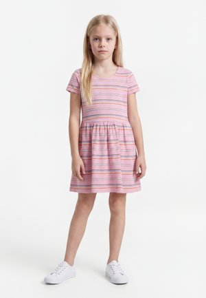 Jeune fille debout portant une robe rose à manches courtes avec des rayures horizontales et des baskets blanches sur fond blanc.