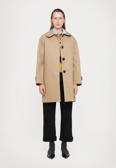Una donna indossa un cappotto beige con bottoni sopra un maglione colorato e una camicia blu, abbinato a pantaloni neri con risvolto e scarpe nere a punta con calzini grigi.