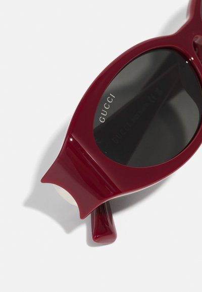 Burgundy-lasit aurinkolasit mustilla linsseillä, joissa on ainutlaatuinen kissansilmämuoto ja kiiltävä pinta. Merkin "GUCCI" logo on kaiverrettu kehykseen.