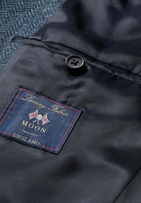 Tessuto navy con un motivo testurizzato, dotato di un bottone nero e un'etichetta con accenti rossi che riporta il testo "Luxury Fabric MOON England."