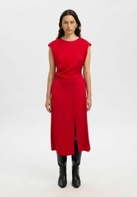 Frau mit langen dunklen Haaren, die ein ärmelloses rotes Midikleid mit einem Schlitz vorne und schwarzen Knielangen Stiefeln trägt, steht vor einem hellen Hintergrund.