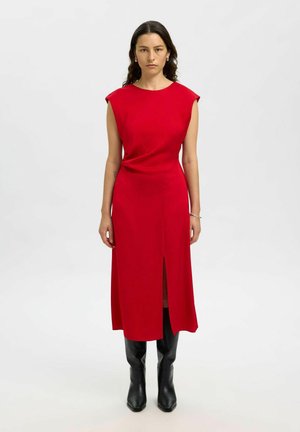 Vrouw met lang, donker haar, gekleed in een mouwloze rode midi-jurk met een split aan de voorkant en zwarte kniehoge laarzen, staande op een lichte achtergrond.