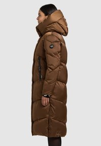 Dolga rjava puhasta jakna z visokim ovratnikom, žepki z zadrgo in prešito teksturo. Na zgornjem delu roke je logotip, ima pa tudi strukturiran kapuco.