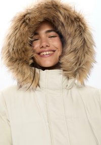 Krémová parka s kapucí podšitou kožešinou, která se vyznačuje béžovými a hnědými tóny. Materiál vypadá hladce a má prošívané detaily.