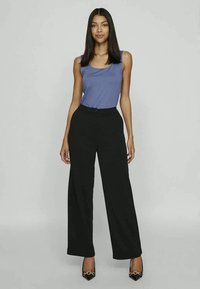 Mulher com cabelo preto longo, vestindo uma blusa sem mangas azul, calças largas pretas de cintura alta e sapatos de salto pretos pontiagudos com detalhe em corrente dourada.
