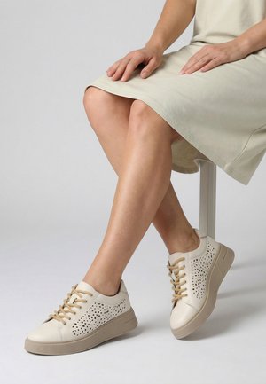 Ben krydset, mens de sidder, iført beige perforerede sneakers med tan snørebånd og en lysebeige kjole, mod en ens grå baggrund.