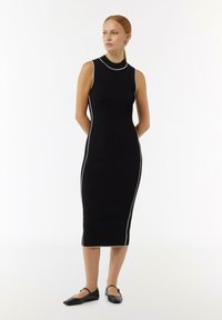 Schwarzes geripptes Midikleid mit hohem Ausschnitt, das weiße Seitenpipings aufweist. Das Kleid hat eine taillierte Silhouette und wird mit schwarzen Ballerinas kombiniert.