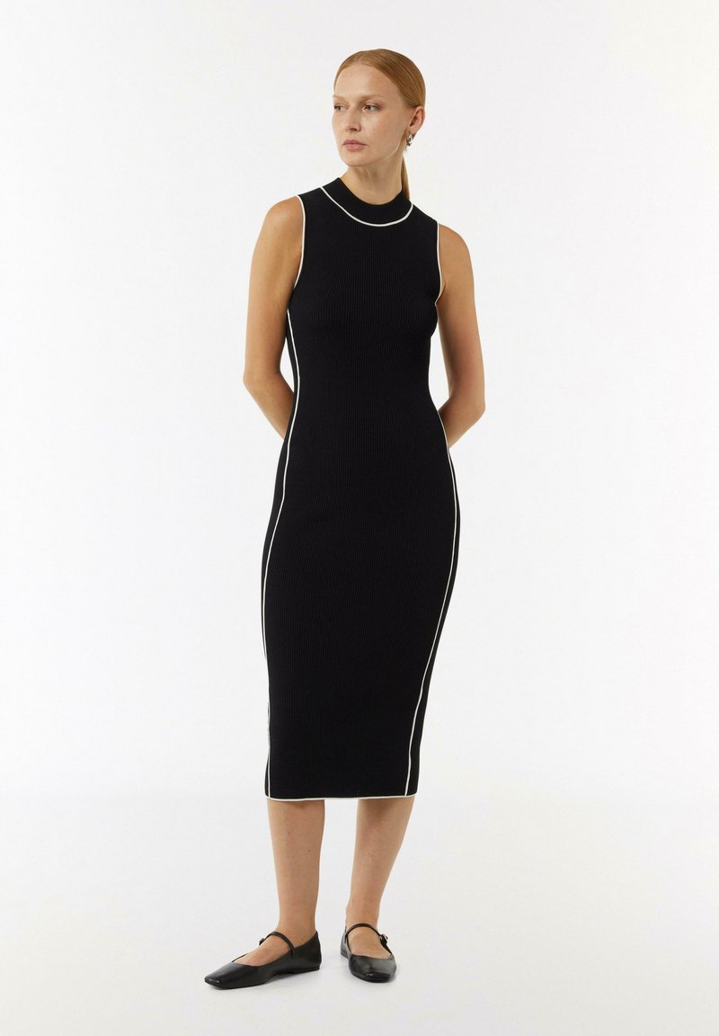 Schwarzes geripptes Midikleid mit hohem Ausschnitt, das weiße Seitenpipings aufweist. Das Kleid hat eine taillierte Silhouette und wird mit schwarzen Ballerinas kombiniert.