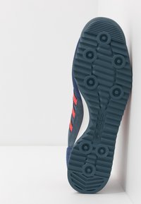 Granatowy zamszowy sneaker z czerwonymi paskami, białą podeszwą środkową oraz teksturowaną gumową podeszwą z okrągłymi wzorami bieżnika i oznaczeniami marki.