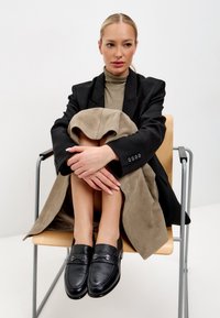 Černé kožené mokasíny s dekorativní kovovou sponou, zkombinované s béžovým semišovým kabátem, který je přehozený přes zkřížené nohy na minimalistické židli.