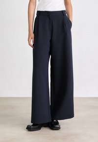 Pantaloni a gamba larga color navy realizzati in tessuto testurizzato, con una sagomatura a pieghe frontali, vita alta e tasche laterali. Abbinati a scarpe nere.