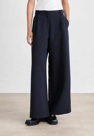 Pantaloni a gamba larga color navy realizzati in tessuto testurizzato, con una sagomatura a pieghe frontali, vita alta e tasche laterali. Abbinati a scarpe nere.
