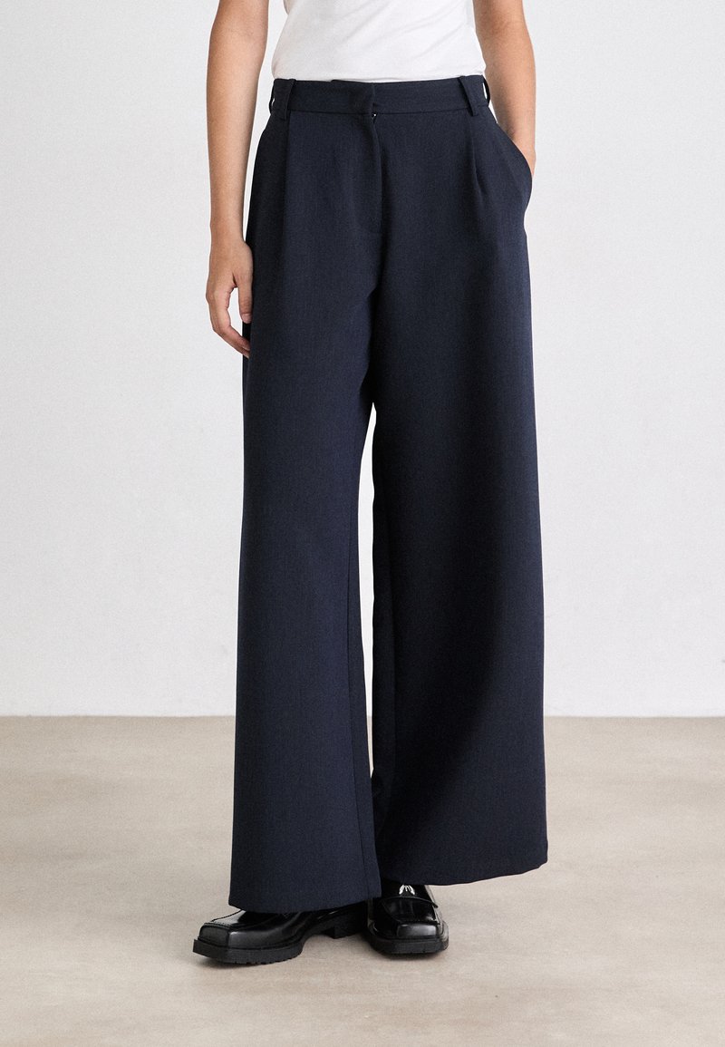 Pantaloni a gamba larga color navy realizzati in tessuto testurizzato, con una sagomatura a pieghe frontali, vita alta e tasche laterali. Abbinati a scarpe nere.