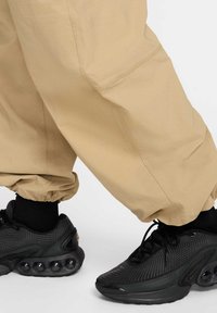 Pantalons cargo beiges avec des poignets élastiques, associés à des baskets noires avec une tige texturée et des éléments de coussinage à air prononcés.