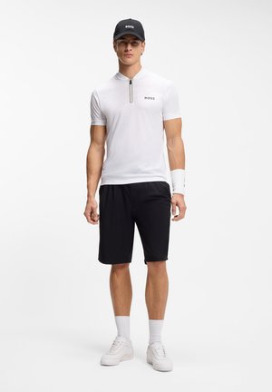 Hombre con gorra negra, camisa blanca de manga corta con cremallera, pantalones cortos negros, muñequeras blancas, calcetines y zapatillas deportivas, de pie contra un fondo blanco liso.