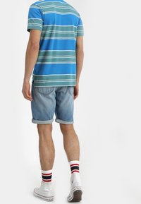 Randig blå och grön kortärmad skjorta, ljusa denimshorts, vita högtoppsskor med röd, vit och blå randiga strumpor.