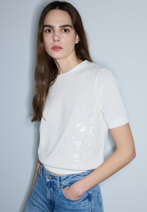 Mujer con cabello largo y castaño, con un top de punto blanco de manga corta con sutiles lentejuelas y vaqueros azules, posando con una mano en el bolsillo.