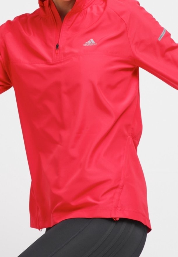 Chaqueta roja de Adidas con media cremallera, usada por una persona con leggings negros, mostrando detalles frontales y laterales, logo en el pecho y tira reflectante en la manga.