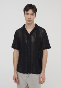 Camicetta nera a maniche corte con un design a maglia aperta e testurizzata, caratterizzata da strisce verticali e colletto. Chiusura con bottoni e vestibilità rilassata.