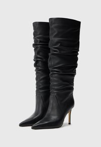 Ein Paar schwarze, lässige, knielange Lederstiefel mit spitzem Zeh und Stiletto-Absätzen auf einem schlichten Hintergrund.