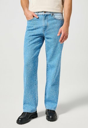 Mann trägt hellblaue, strukturierte Jeans mit geradem Bein, schwarze klobige Schuhe und ein beiges Hemd, steht mit einer Hand in der Tasche.