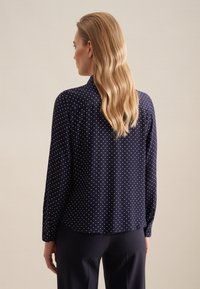 Blusa a maniche lunghe blu navy con pois bianchi, dettaglio colletto e polsini con bottoni, realizzata in tessuto leggero con una consistenza liscia.