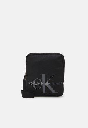 Sac bandoulière carré noir avec logo gris "cK Calvin Klein Jeans" et bandoulière réglable, fermeture zippée sur le dessus.
