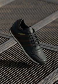 Svarta Adidas-sneakers med bruna ränder och gult logo på en metallgitteryta, vinklade för att visa detaljer från topp och sida.
