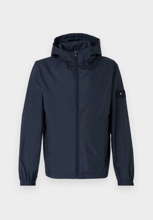Marineblauwe waterdichte jas met capuchon, elastische manchetten, een rits aan de voorkant en een borstzak met een logo. Glad textuur en lichtgewicht ontwerp.