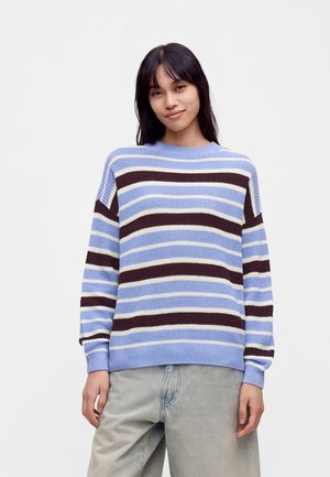 Ung person med langt sort hår iklædt en blå, hvid og mørkebrun stribet sweater og lysegrå bukser, stående mod en ensfarvet baggrund.