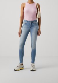 Jeans skinny de color azul claro con un degradado desvanecido, combinados con una camiseta de tirantes rosa de canalé y zapatillas de color neutro. Se lleva un pequeño bolso blanco de hombro.