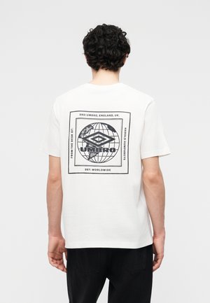 Man with curly dark hair wears a white t-shirt with a black Umbro logo and globe graphic printed on the back, paired with black pants.