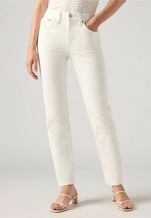 Femme portant un jean slim blanc taille haute et des sandales à talons beiges à brides, debout avec une main dans la poche.