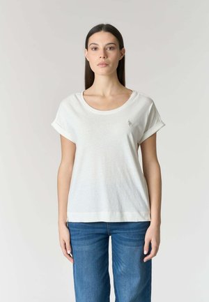 Donna in piedi di fronte, indossa una maglietta bianca a maniche corte con risvolti e jeans blu, su sfondo neutro.