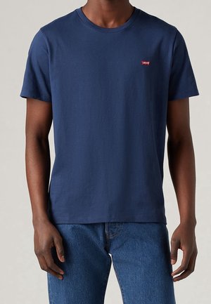 Mand iført en ensfarvet mørkeblå T-shirt med korte ærmer og blå jeans, stående foran en neutral baggrund.