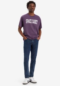 Camiseta de algodón morada con "SPACE CADET" impreso en blanco, combinada con jeans de mezclilla azules y zapatillas en blanco y negro.