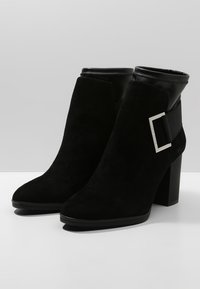 Stivaletti in suede nero alla caviglia con punta affusolata, tacco chunky e polsino in pelle nera liscia. Presentano un accento con fibbia quadrata argentata.